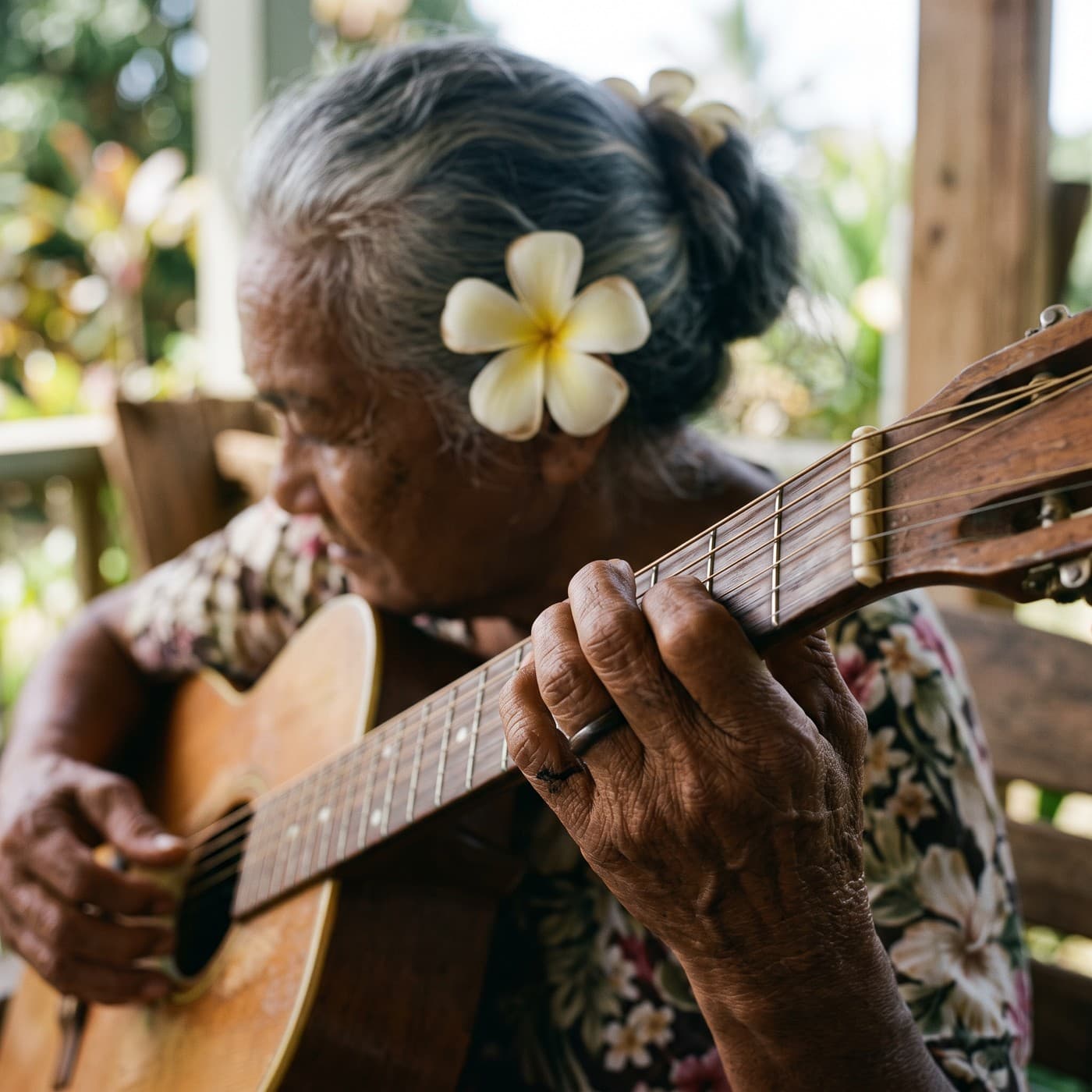 Close view of Auntie Mapuana's hands on the neck of an old acoustic guitar, a plumeria in her gray hair